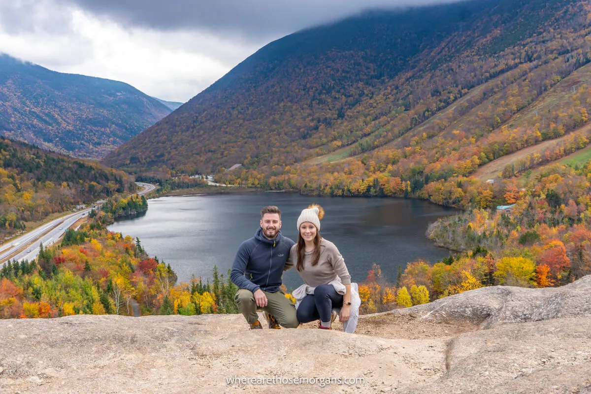 Two hikers crouching down for a photo together on a rocky outcrop overlooking a lake and hill slopes covered in trees on a cloudy day in New England