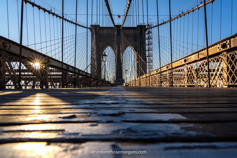 Empty Brooklyn Bridge pedestrian walkway with the sun peaking through a gap in the metal fence at sunrise in NYC