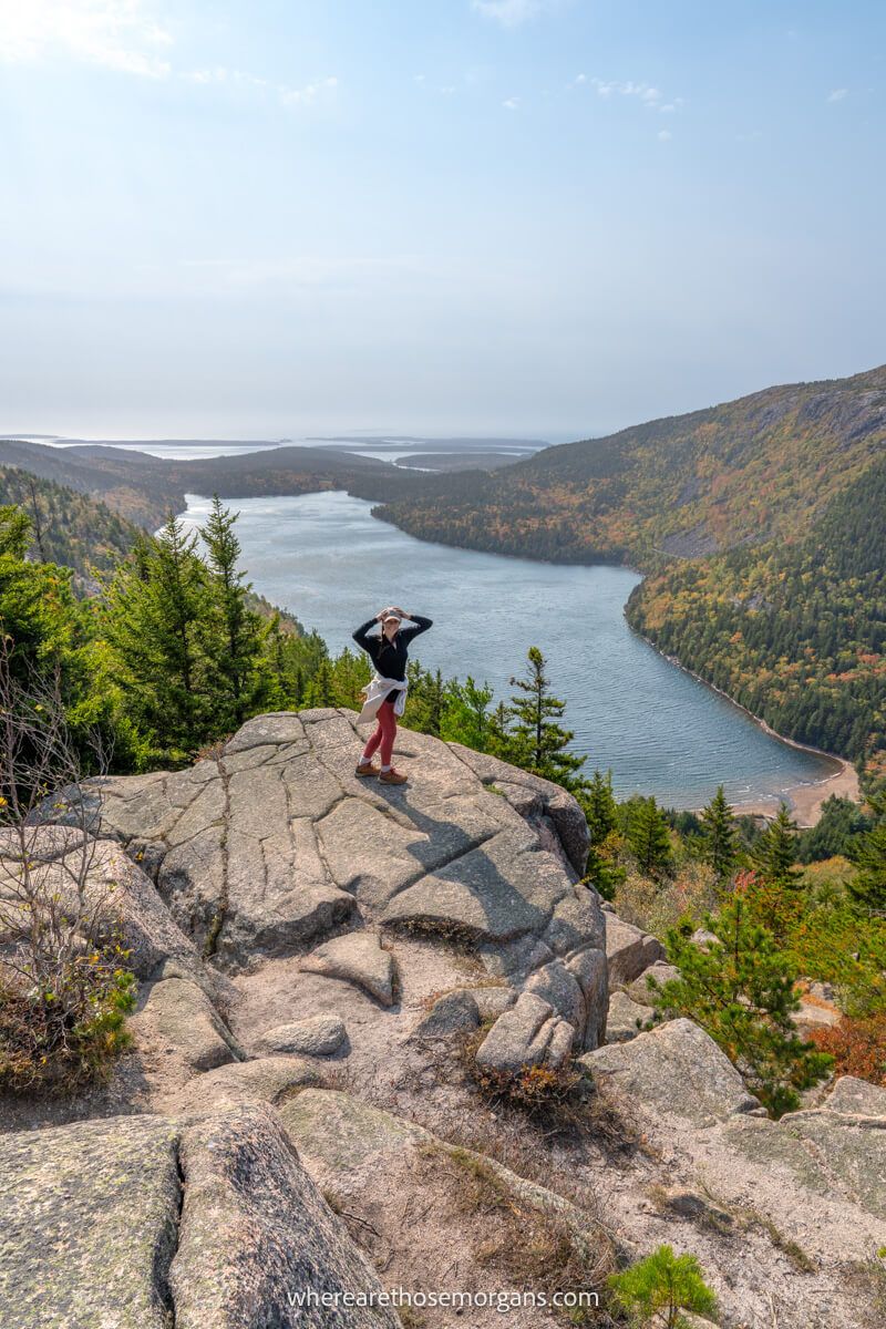 Photo of a hiker stood on a rocky ledge in the distance holding a hat down from blowing away with far reaching views over a lake and rolling hills with trees