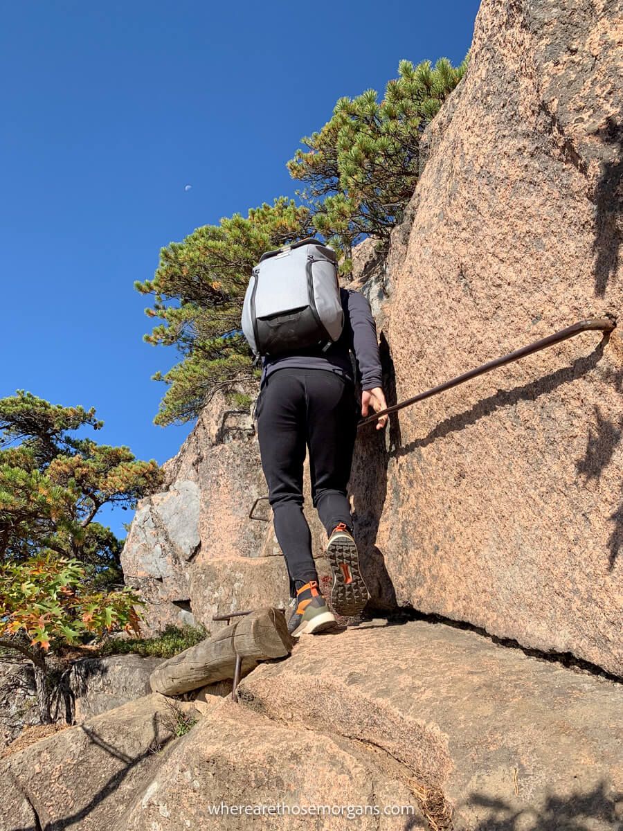 Hiker climbing a narrow rock path holding onto metal bars for balance