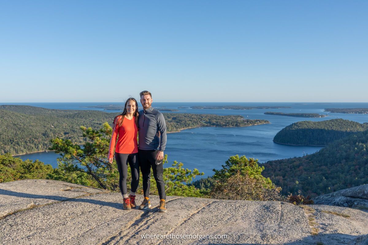 Two hikers stood together on a rocky hiking trail summit in Acadia National Park with views over lakes, trees and the ocean behind