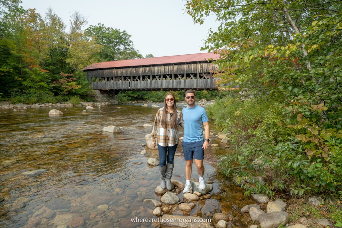 Couple standing together on rocks at the side of a river with trees and a covered bridge with red roof behind