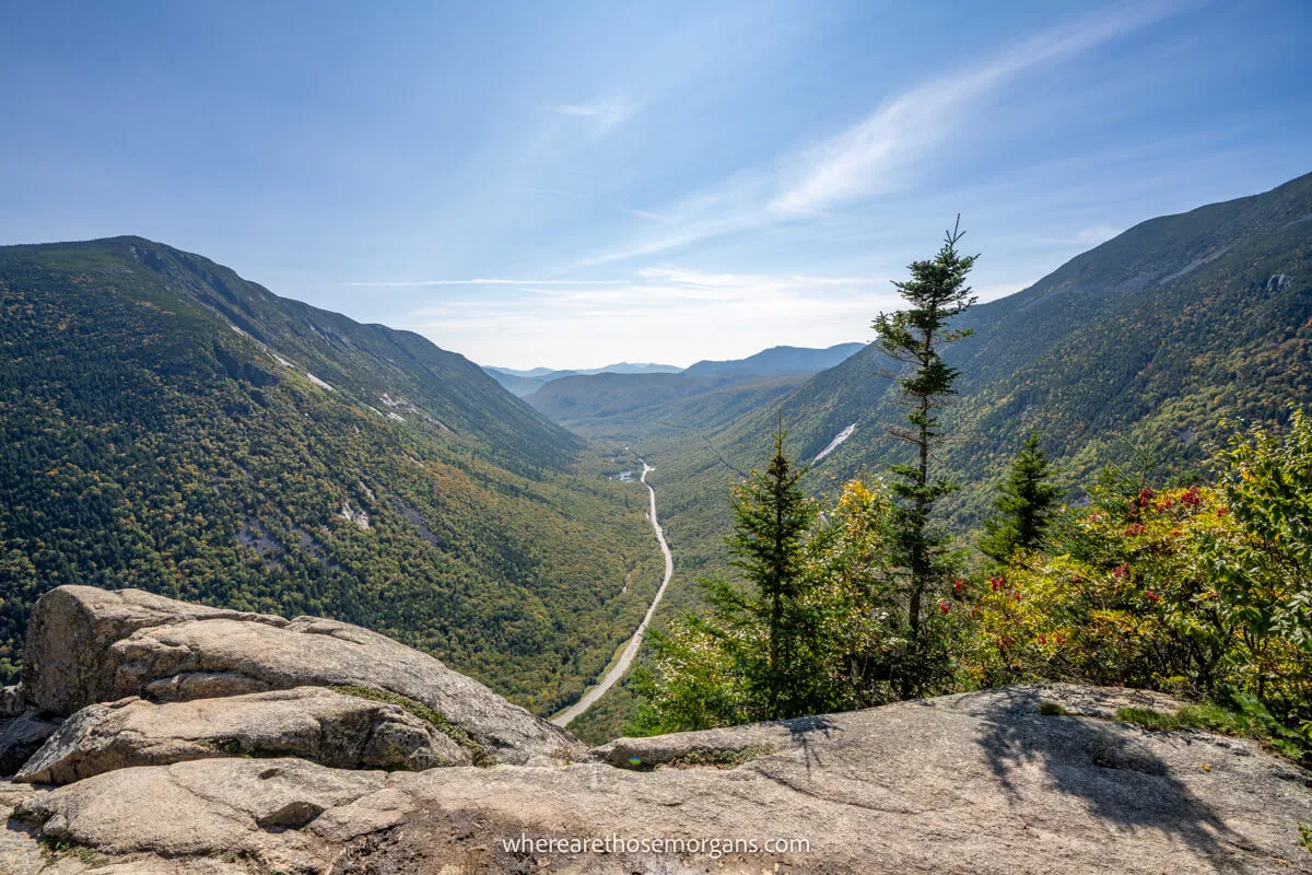 Photo of a rock ledge leading to far reaching views over a narrow road cutting through a canyon filled with trees on a sunny day