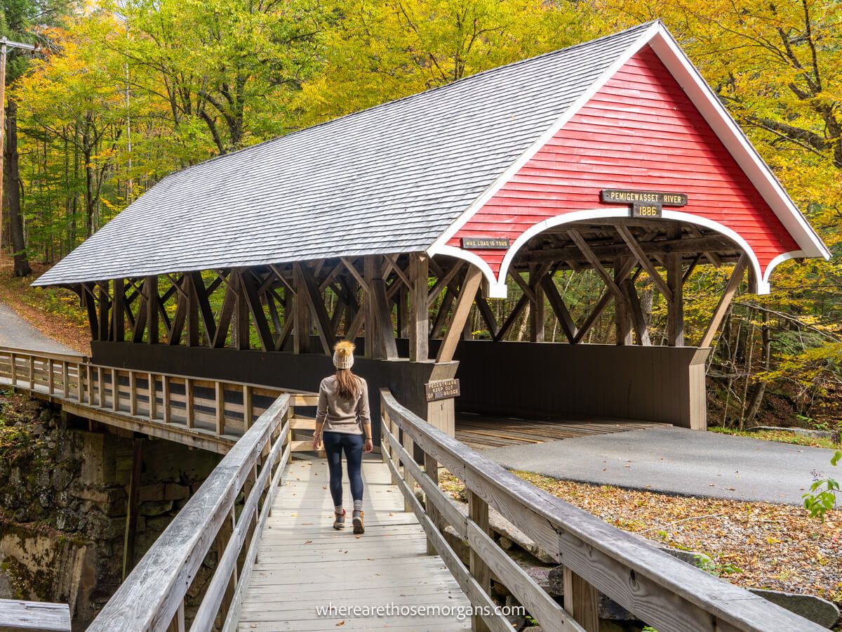 Hiker walking across a wooden bridge to a wooden covered bridge with red paneling on the front and a triangular shaped roof and yellow leaves in trees behind in the Flume Gorge in New Hampshire