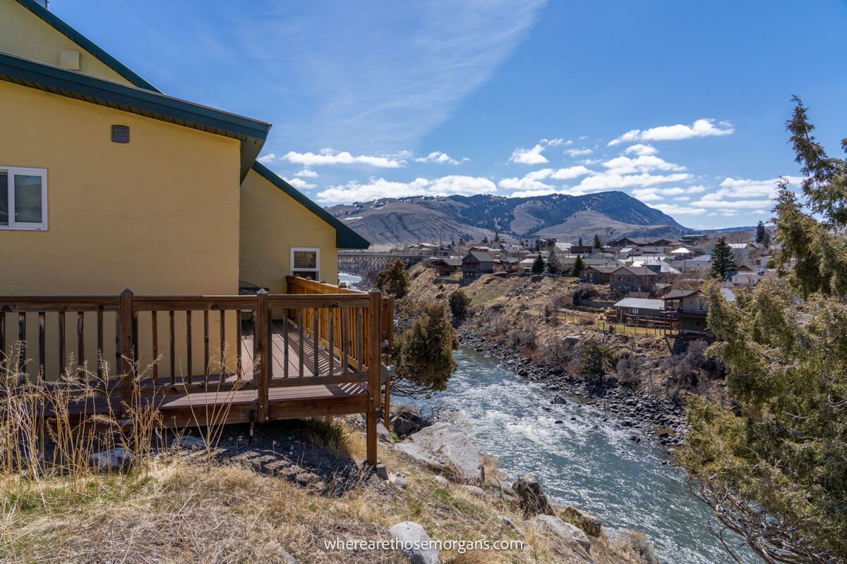 Photo of the side of a yellow cottage with a wooden deck on the side of a bank leading down to a river and hills in the distance in Gardiner MT one of the best places to stay near Yellowstone National Park