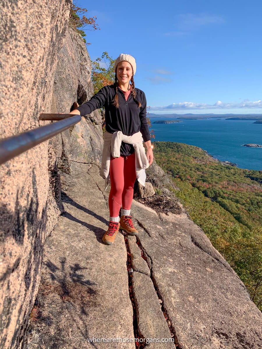 Hiker on a narrow ledge with long drops leading to views over trees and the ocean holding onto an iron rung bar on the Precipice Trail in Acadia