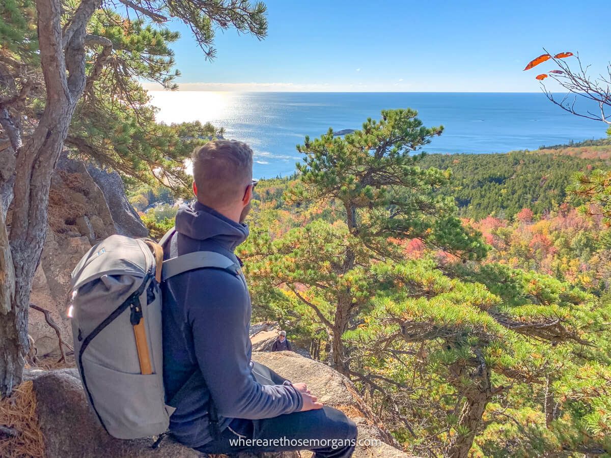 Hiker sat on a narrow rock ledge with a backpack staring out at the ocean and fall foliage colors in nearby trees