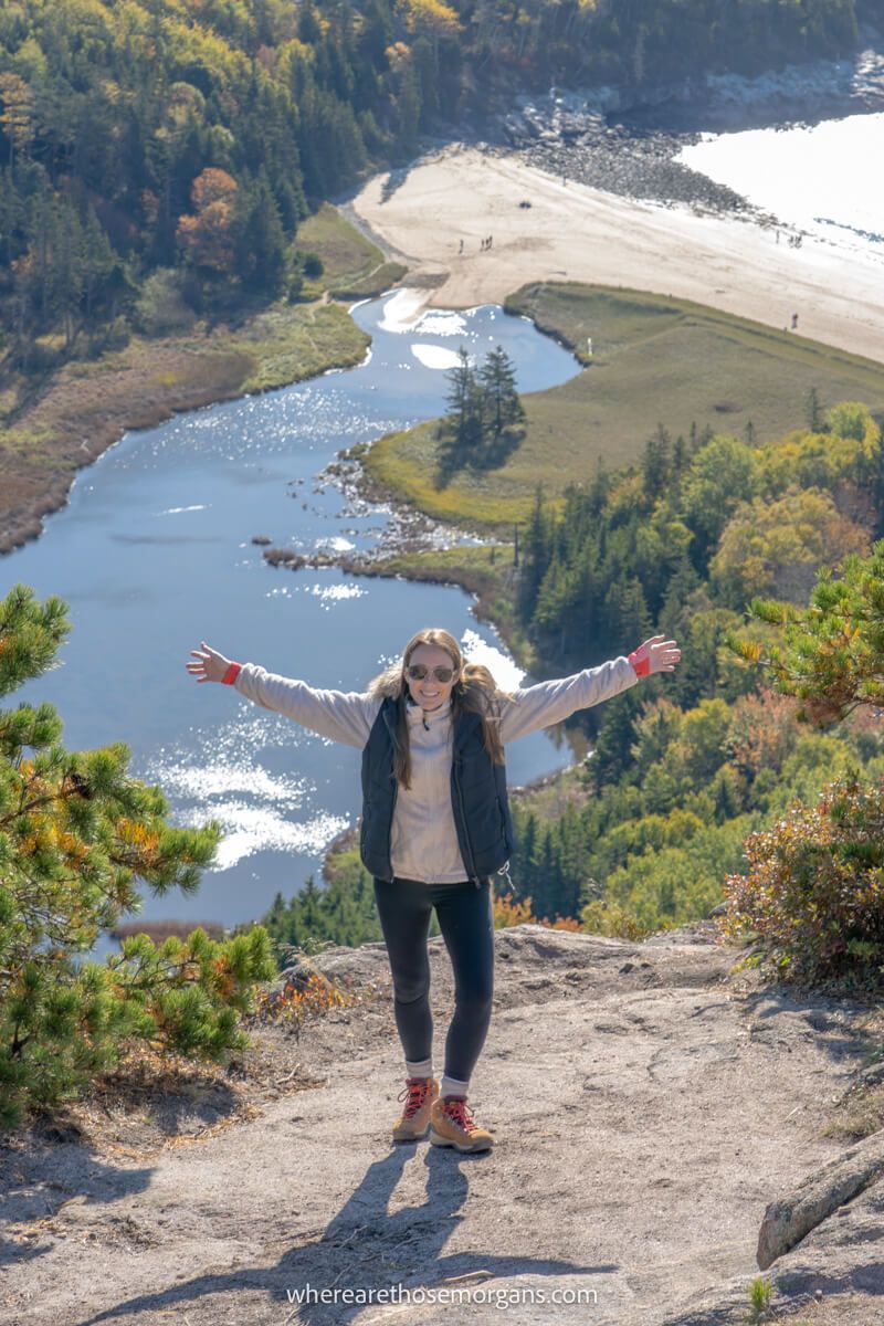 Hiker stood at on a rocky summit with arms outstretched and views over a beach in the distance below