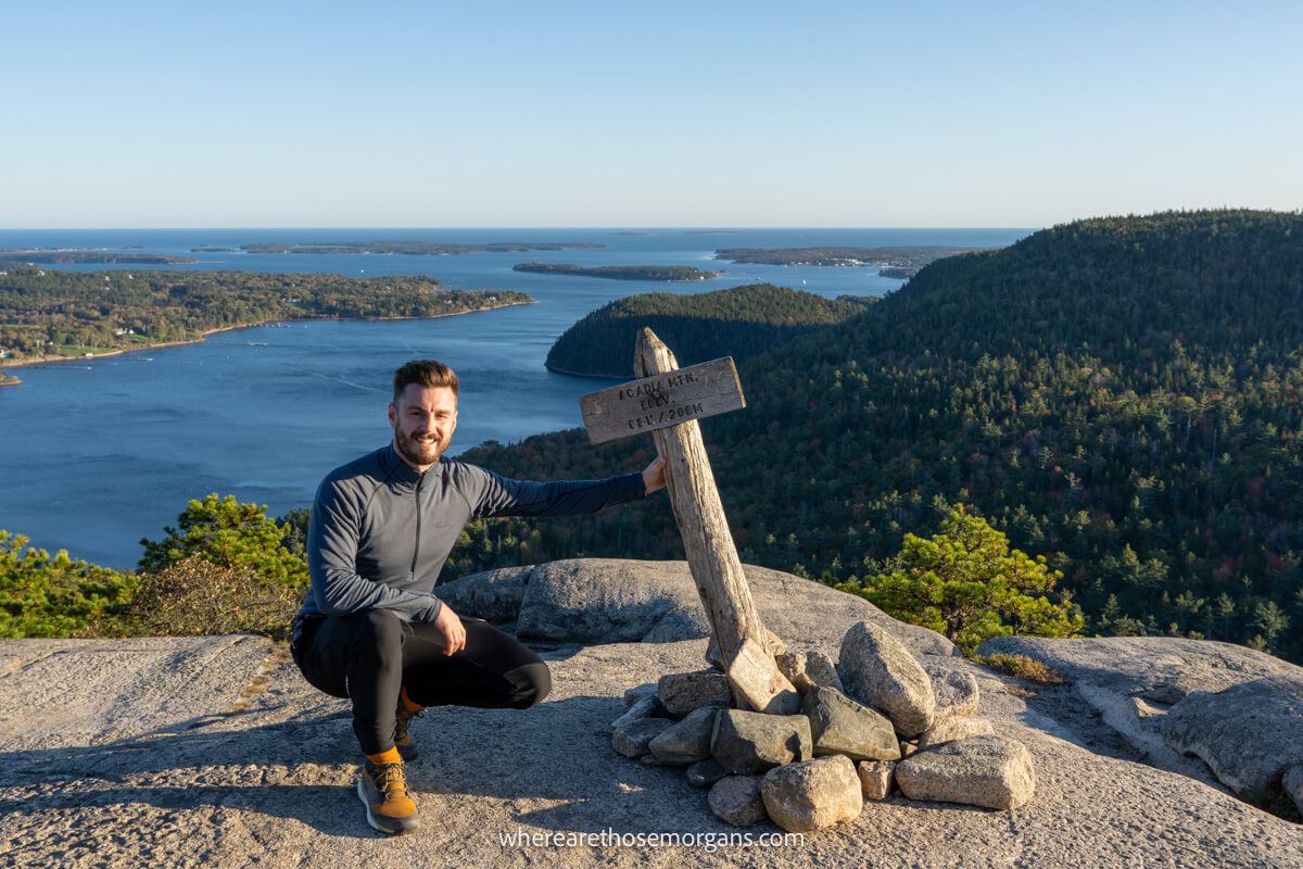 Hiker crouched on a rocky summit next to a small wooden sign with views over lakes, trees and the ocean behind in Acadia National Park