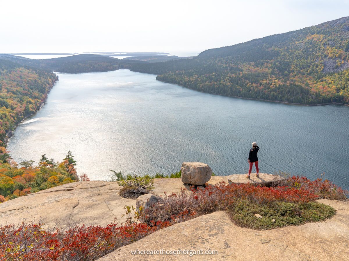 Photo of a hiker in the distance stood on a rock overlooking a large lake and rolling hills covered in trees with the ocean behind on a sunny buy hazy day at Acadia National Park in Maine