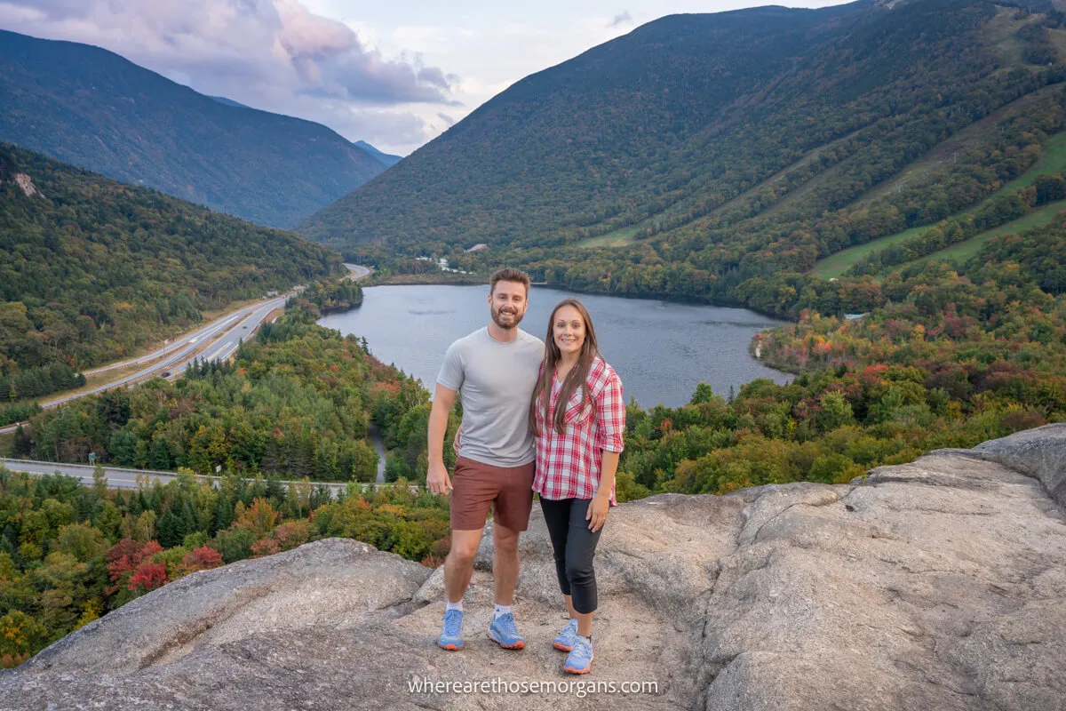 Two hikers standing next to each other on a rocky outcrop with a small lake behind in between hill slopes covered in trees