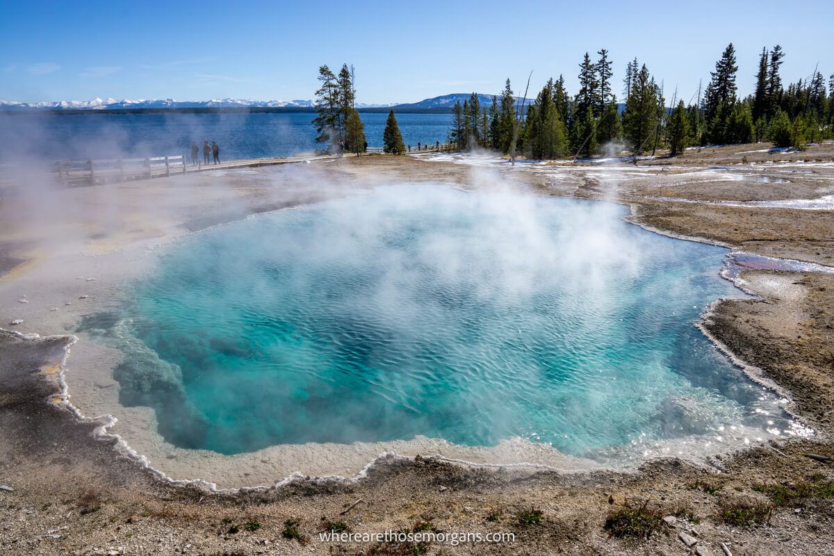 Blue colored hot spring pool with light steam surrounded by yellow grass and a large lake in Yellowstone