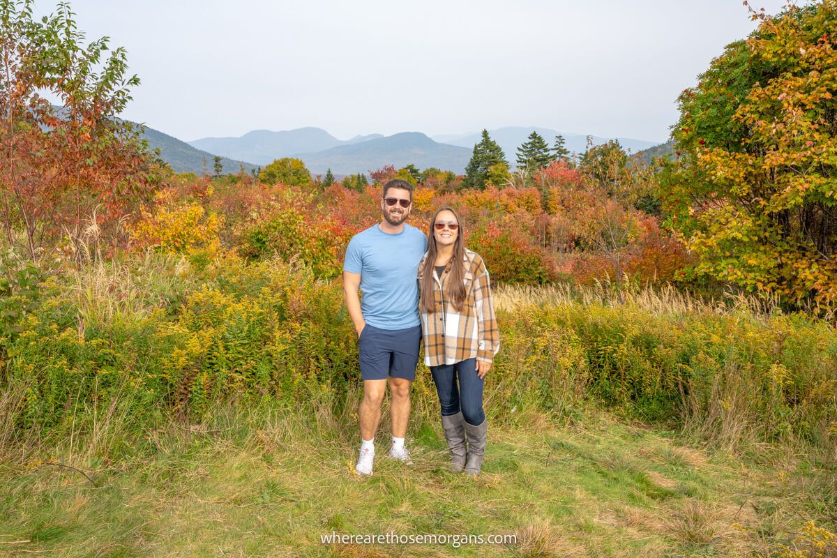 A couple wearing sunglasses in light clothing standing together on grass for a photo with a far reaching view over trees and rolling hills in the background on the Kancamagus Highway with vibrant fall foliage colors