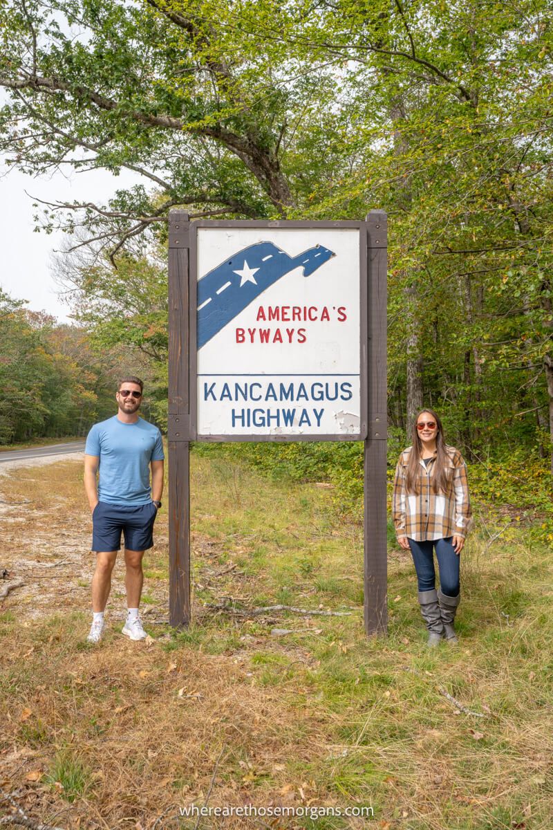 A couple standing on either side of a wooden sign marking the entrance to Kancamagus Highway Scenic Byway in New Hampshire