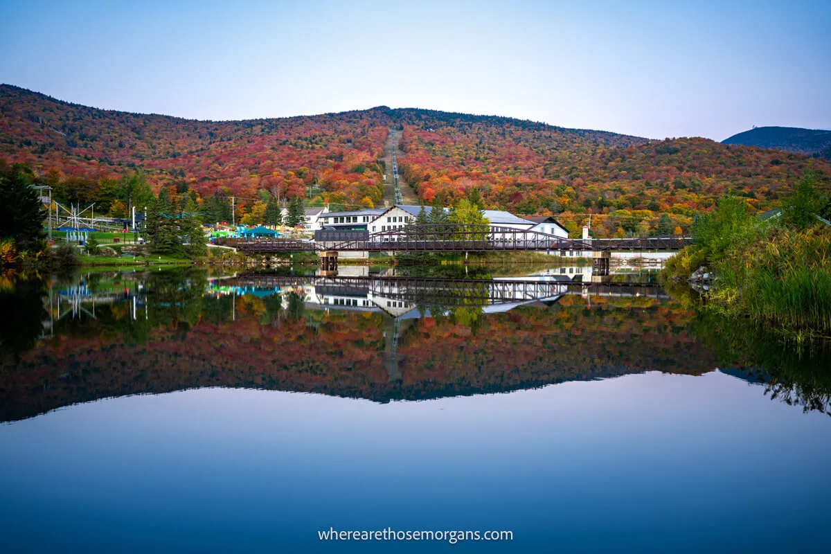 Still lake leading to a reflection of a hillside covered in fall foliage colors and a white building at sunrise in New England