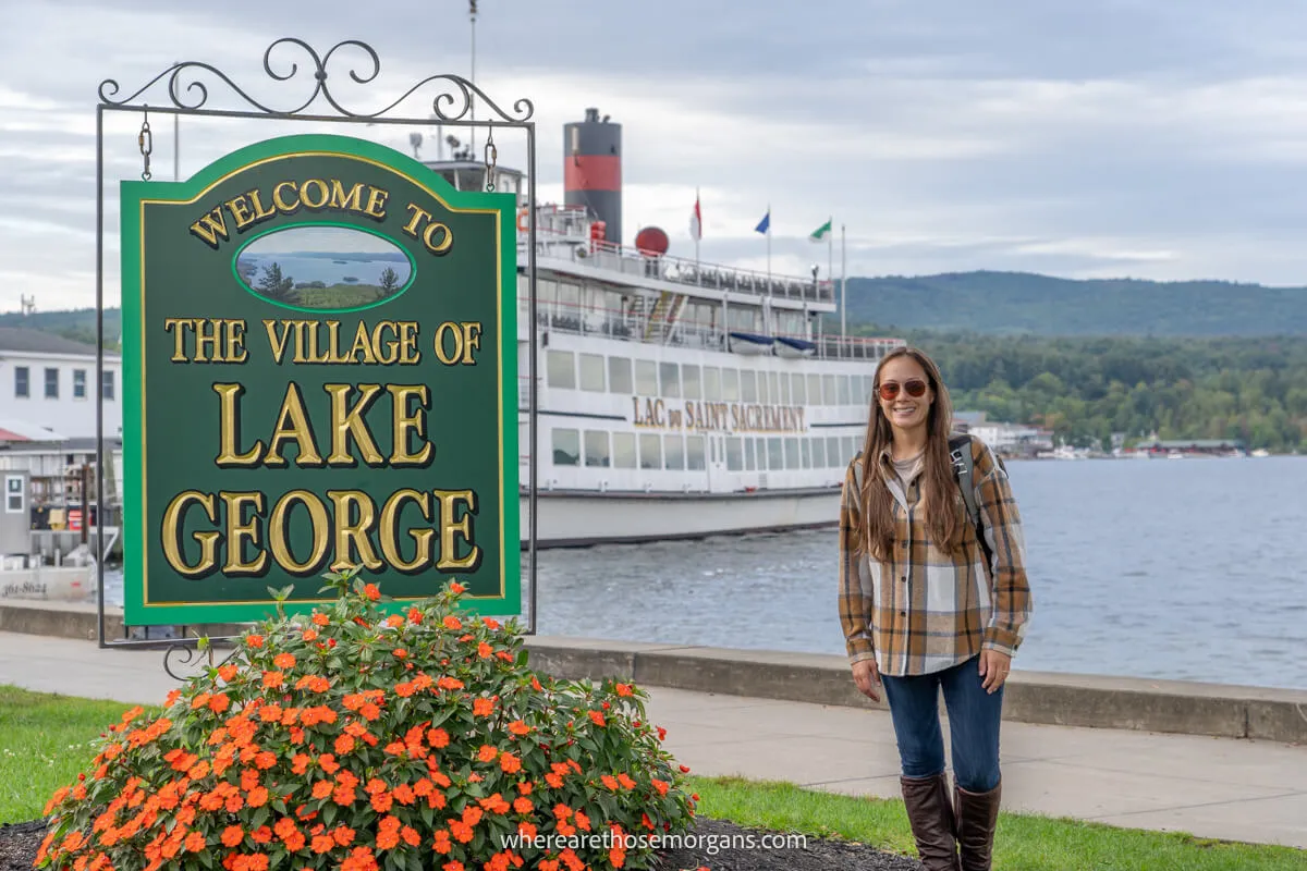 Tourist in a flannel shirt standing next to flowers and a sign for Lake George with a steamboat on a lake behind