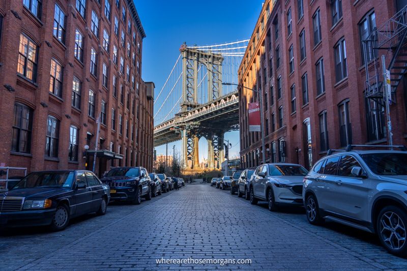 Sunrise view down a street in Brooklyn with cars parked and tall brick buildings leading to a massive bridge