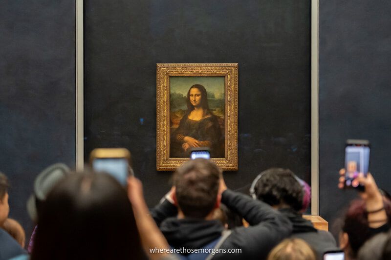 Photo of tourists holding smartphones in the air and all taking photos of the Mona Lisa painting on a dark wall