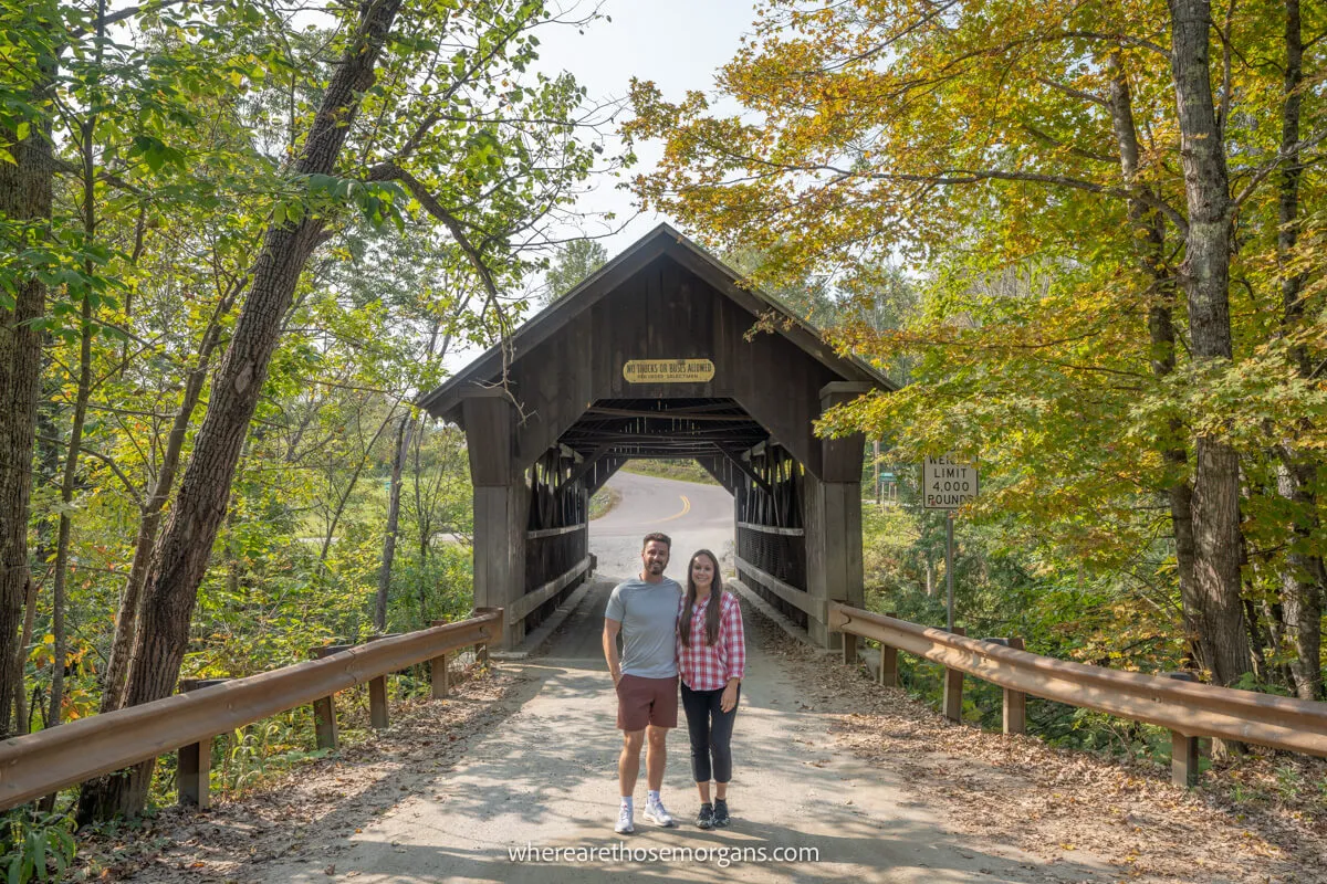 Two tourists stood together on a road for a photo in front of a wooden covered bridge with trees surrounding