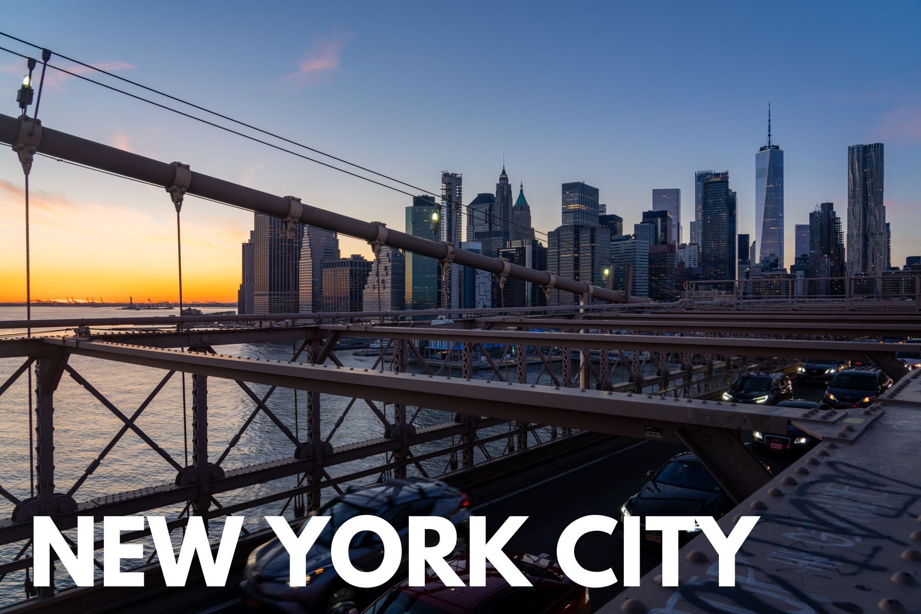 The iconic NYC Lower Manhattan skyline as seen from Brooklyn Bridge at sunset with cars on the bridge below and orange colors above the Statue of Liberty in the distance