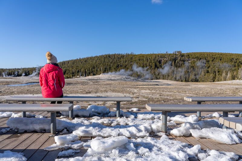 Tourist in red coat and wooly hat sat on a bench alone watching a geothermal field