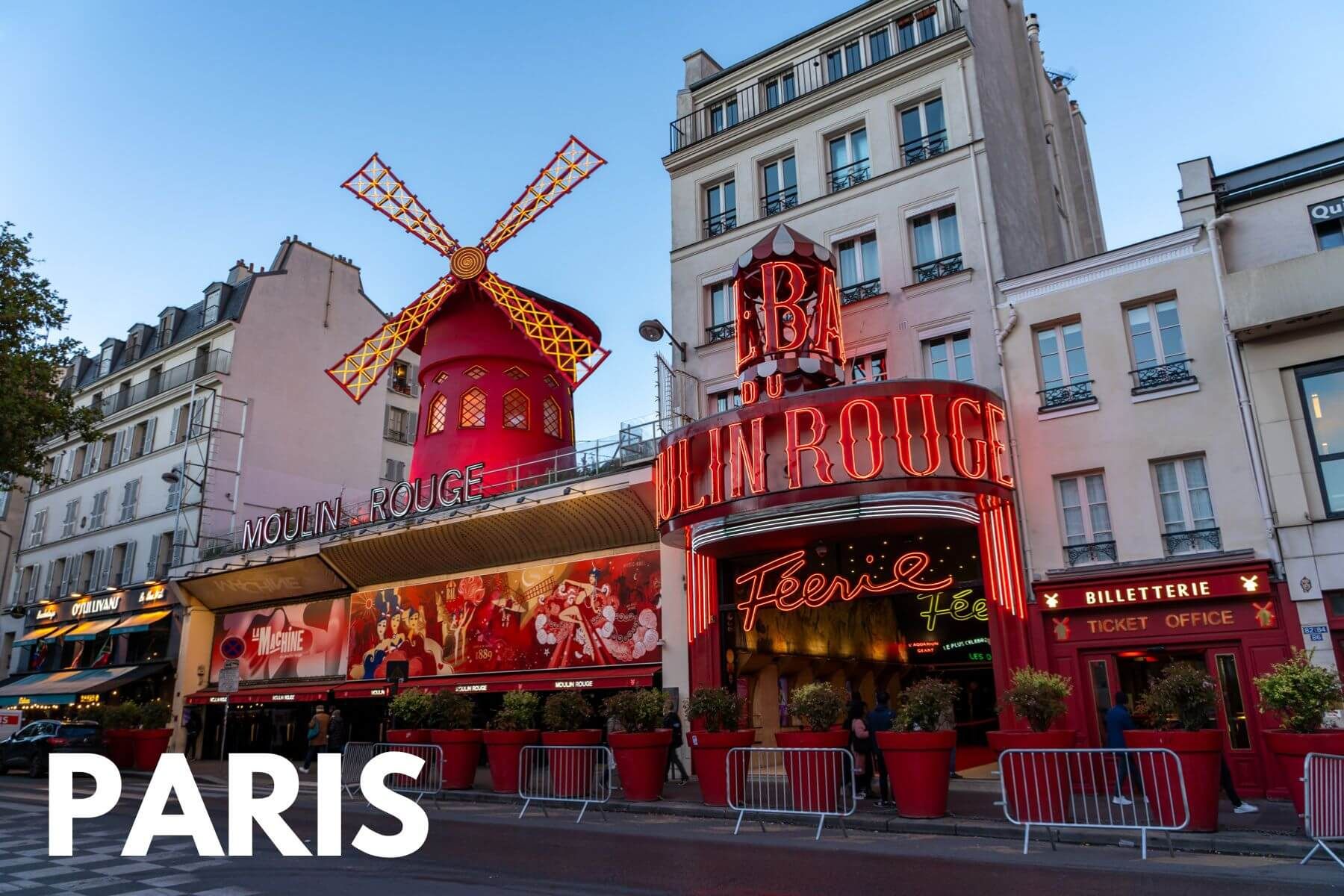 Photo of Moulin Rouge in Paris France at dusk so the windmill and entrance facade are lit up red against a soft blue sky with the Paris overlaid in the bottom left corner