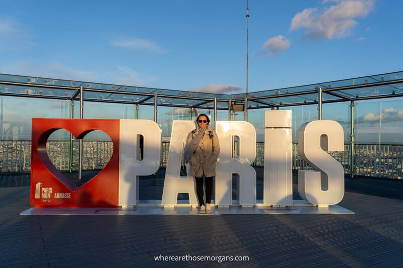 Tourist stood in front of large letters spelling out Paris at sunset on the rooftop of Montparnasse Tower