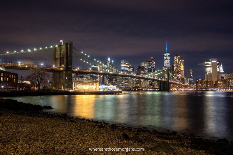 View of Brooklyn Bridge from a pebble beach at night with the city lit up