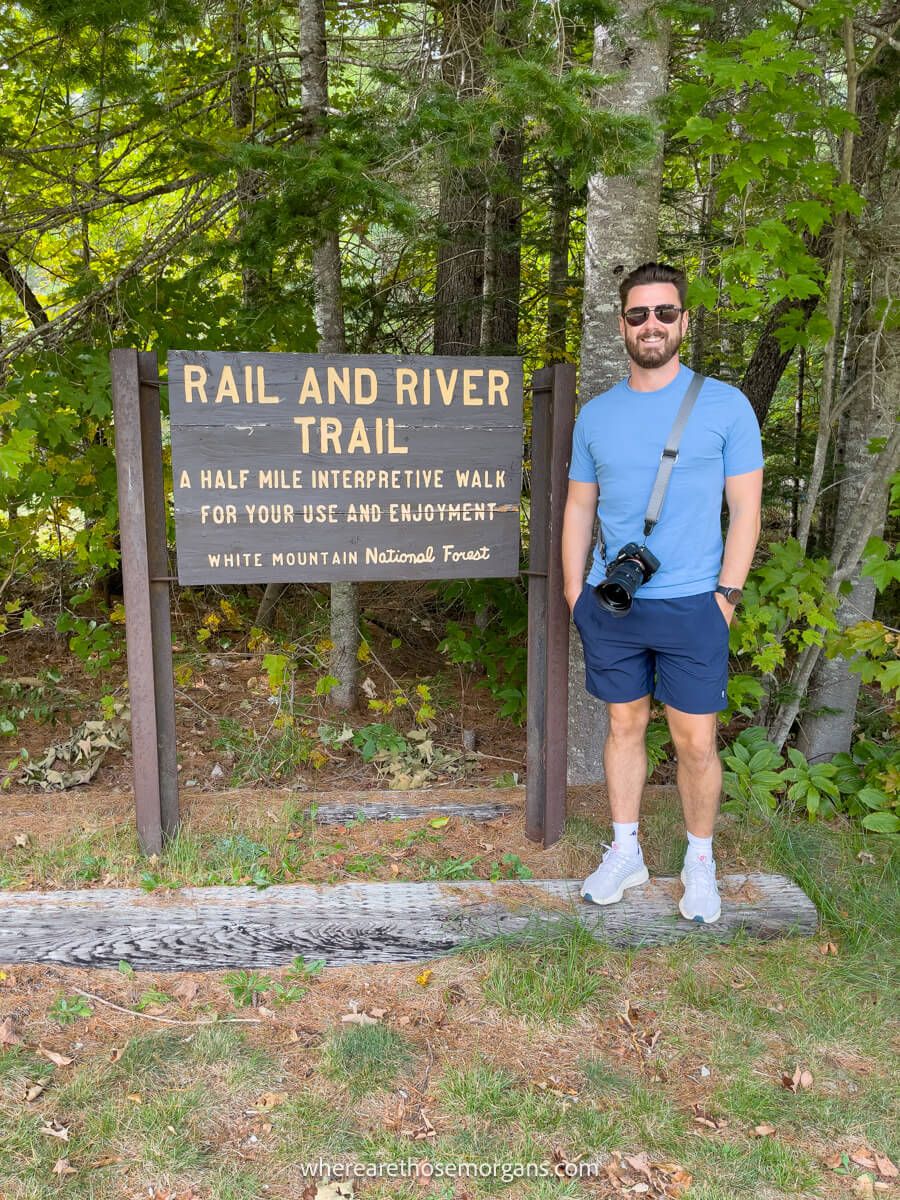 Tourist standing with sunglasses and camera next to a sign marker for a hike in a forest