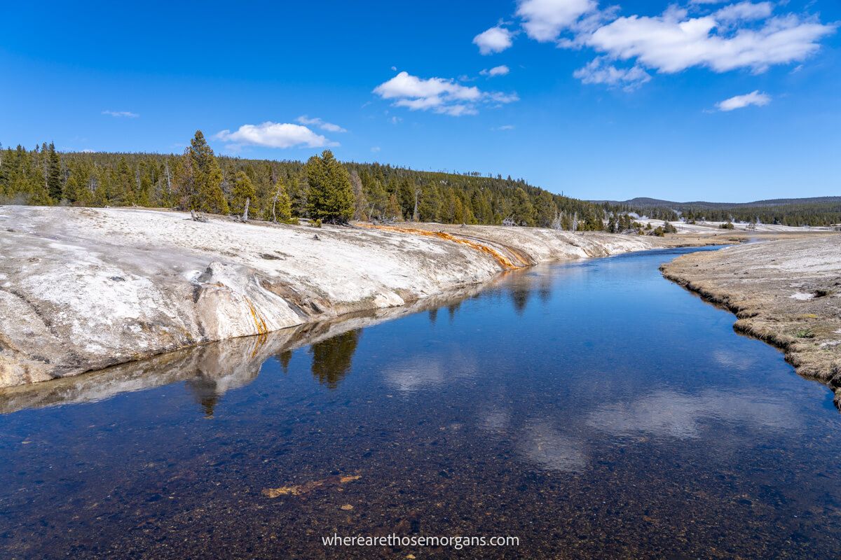 Narrow but perfectly still river with clouds reflecting flanked by geothermal features and trees in Wyoming