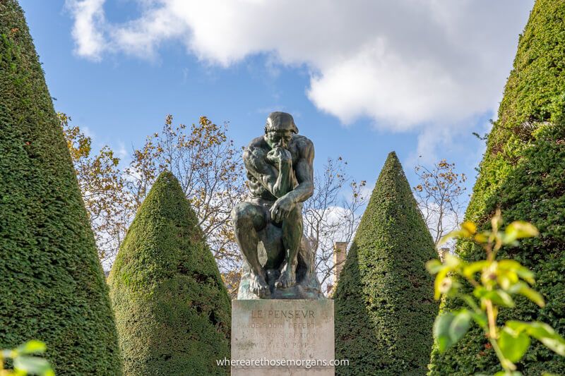 Rodin's The Thinker sculpture in a garden with conical shaped green bushes