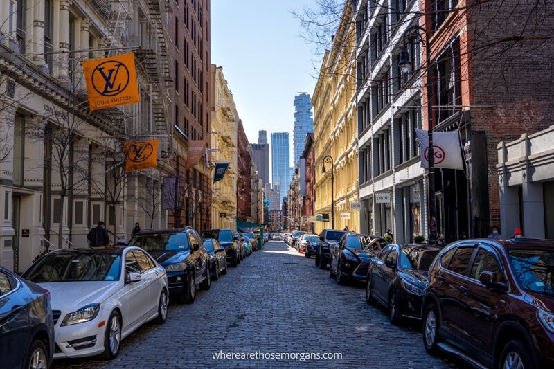 A cobbled street with cars parked on both sides and buildings painted different colors