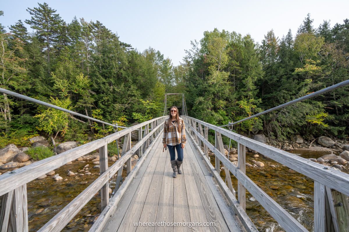 Tourist in plaid shirt and jeans walking across a wooden suspension bridge over a river with trees behind near Lincoln Woods on the Kancamagus Highway in New Hampshire