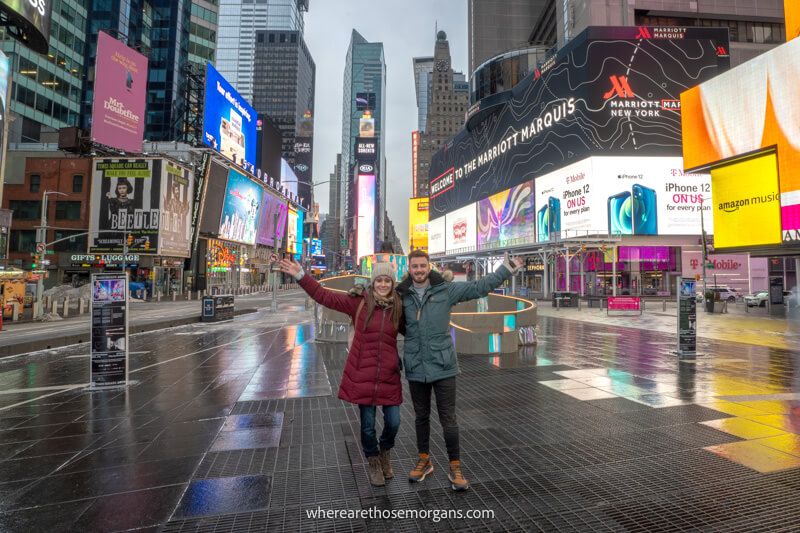 Two tourist stood next to each other with arms in the air as the only people in Times Square NYC with all the lights flashing on boards behind