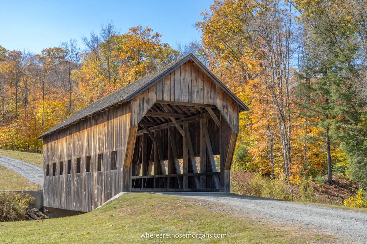 Wooden covered bridge crossing a small creek with trees in the background on a sunny day driving a New England road trip in fall