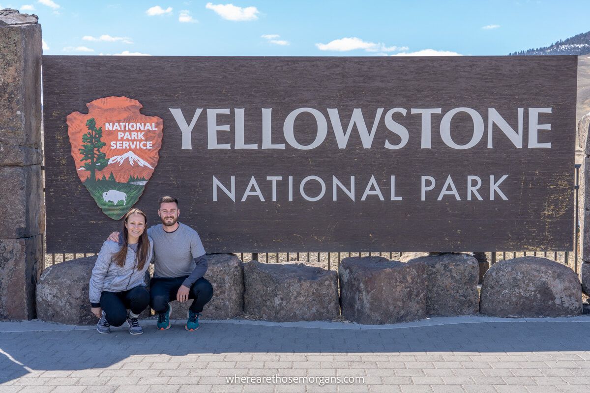 Couple crouched down together in front of the Yellowstone National Park welcome sign on a sunny day