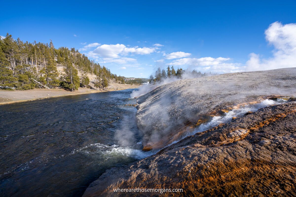 Hot water running down a colorful rocky slope into a river on a sunny day in Yellowstone