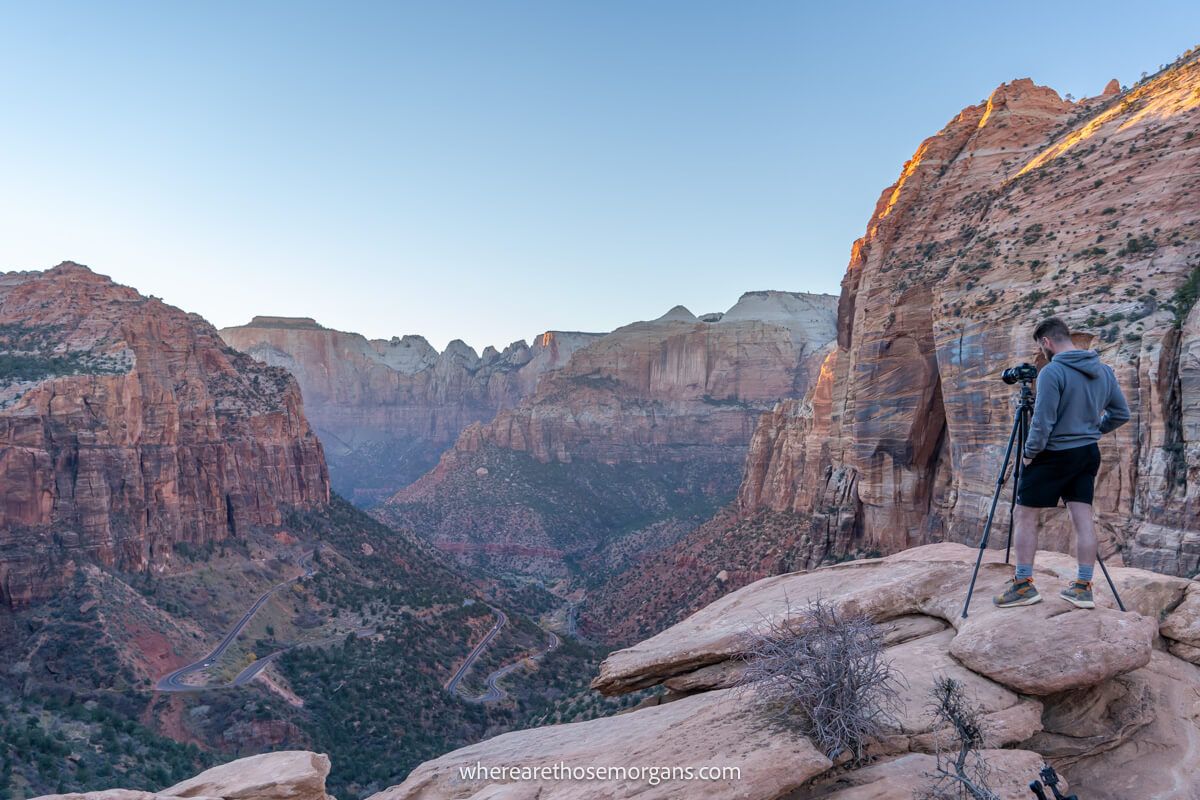 Hiker standing with tripod and camera on a rocky surface overlooking a dramatic canyon and valley view at dusk