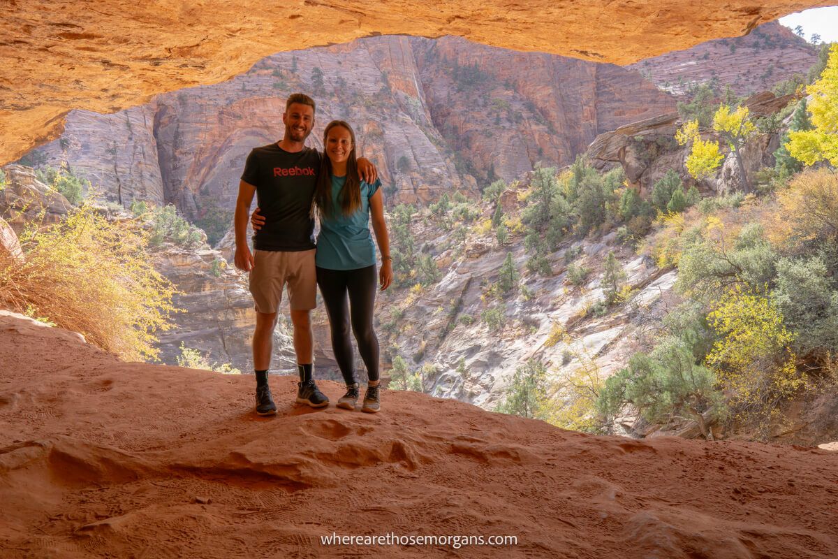 Two hikers stood together for a photo inside a sandstone cave with distant views over a rocky landscape