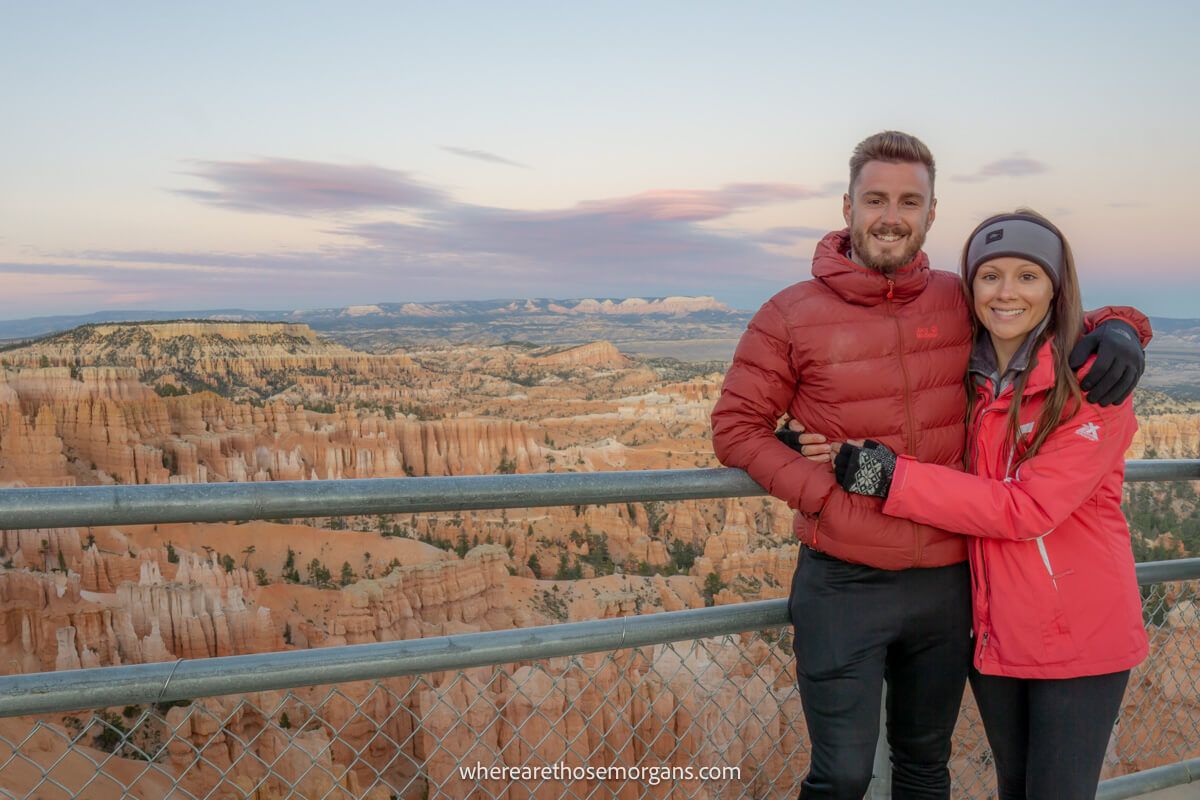 A couple cuddling together in winter clothes next to metal fence with views over a wide open sandstone landscape behind at dusk