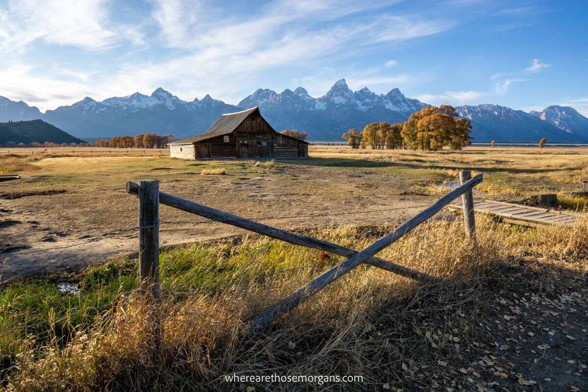 Photo of a small wooden fence with an X-shaped cross in the middle leading to golden yellow meadow, a wooden barn and distant mountains on a sunny day