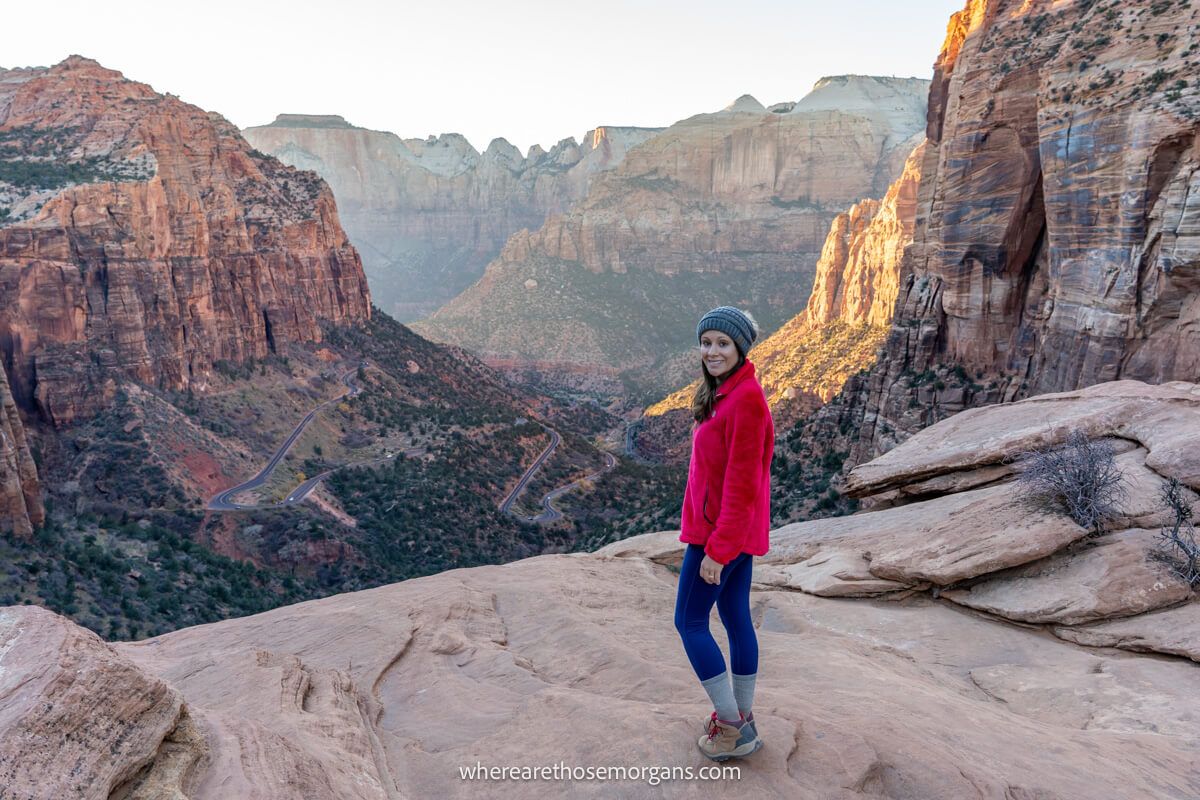 Hiker in red coat and wooly hat standing on the edge of a rocky viewpoint called Zion Canyon Overlook with wide open views of a deep valley with tall cliff walls to either side