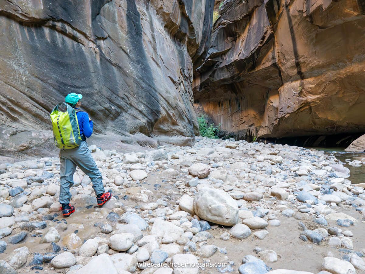 Hiker in waterproof pants and shoes with wooden walking pole standing on small rocks looking at a river slot canyon with tall walls day hiking The Narrows bottom up in Zion