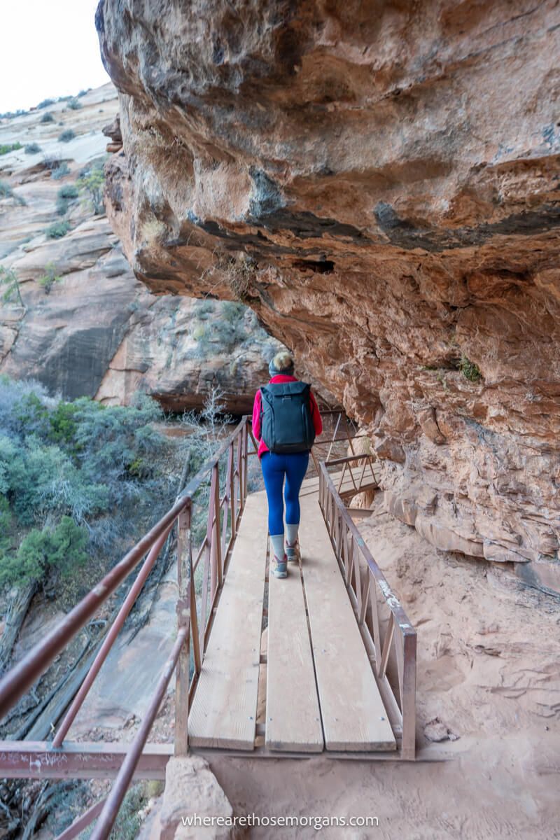 Hiker with backpack in winter clothes walking across a narrow wooden plank next to a red rock cliff wall