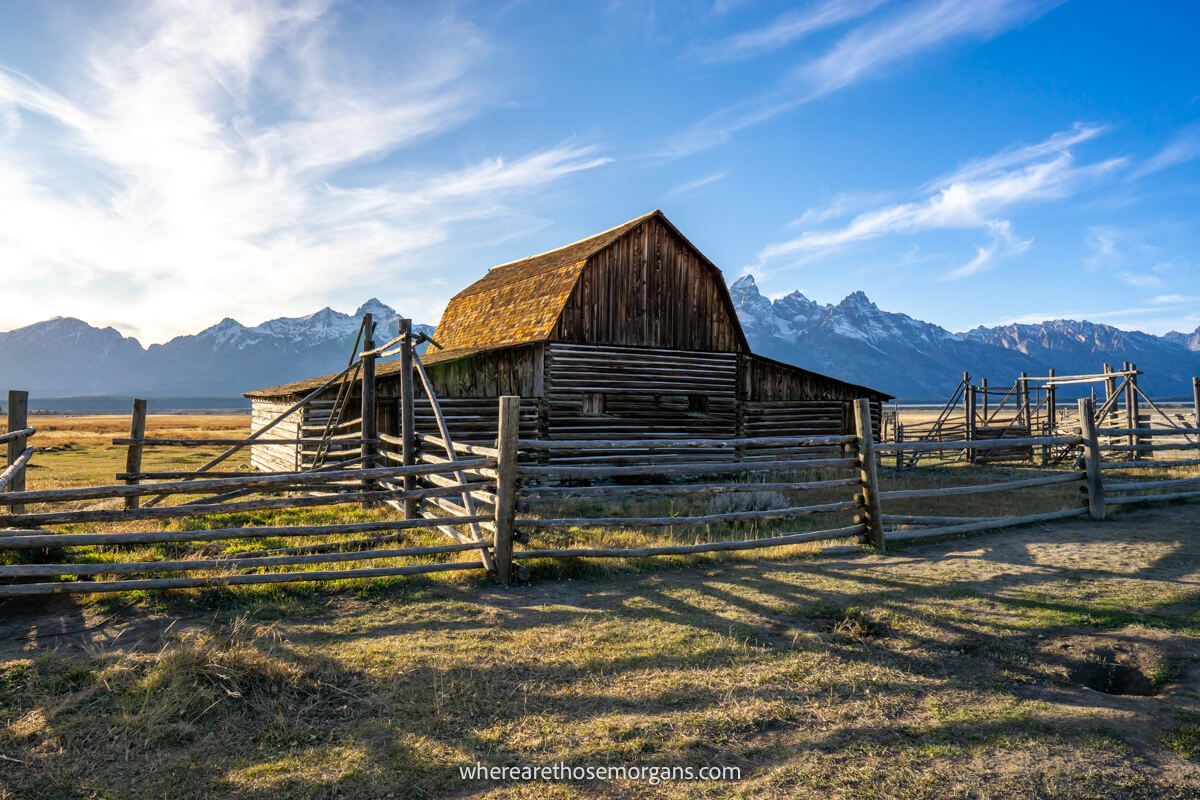 Wide open and flat landscape with a wooden barn surrounded by wooden fencing called John Moulton Homestead leading to distant mountains on a sunny evening