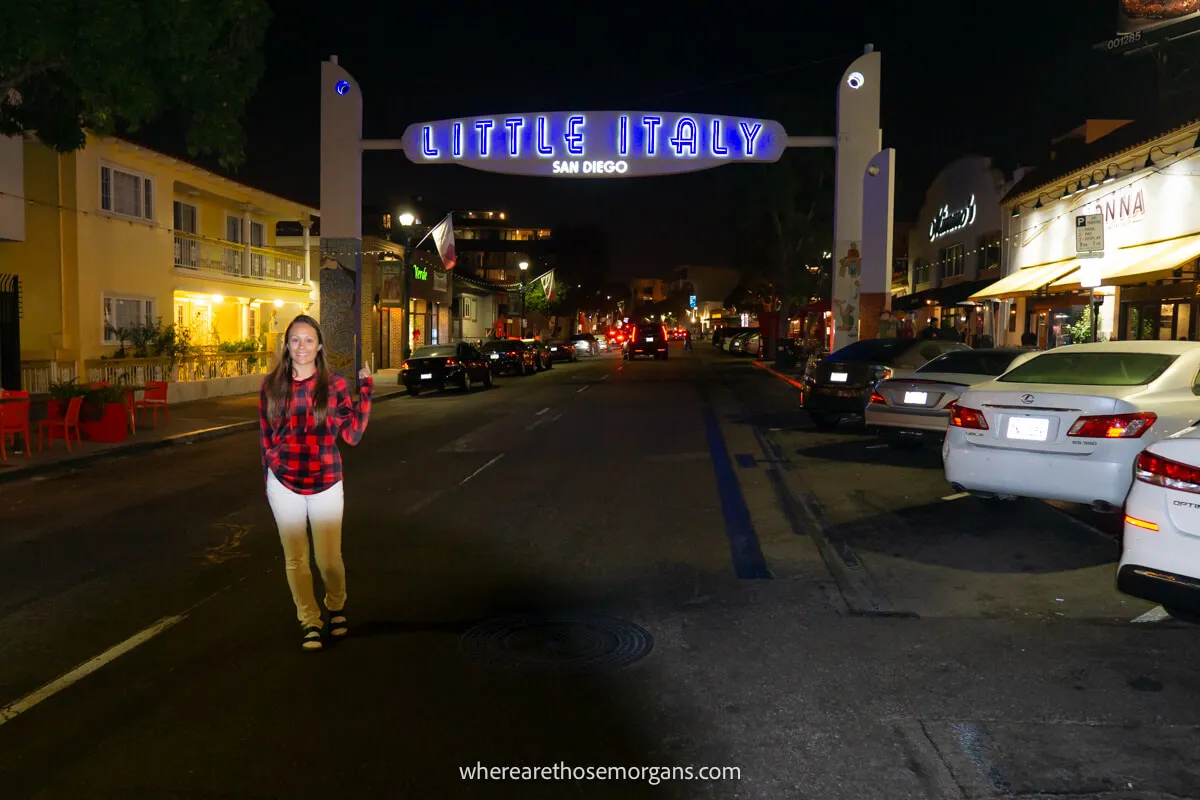 Tourist smiling for a photo on a road with a Little Italy sign behind lit up at night