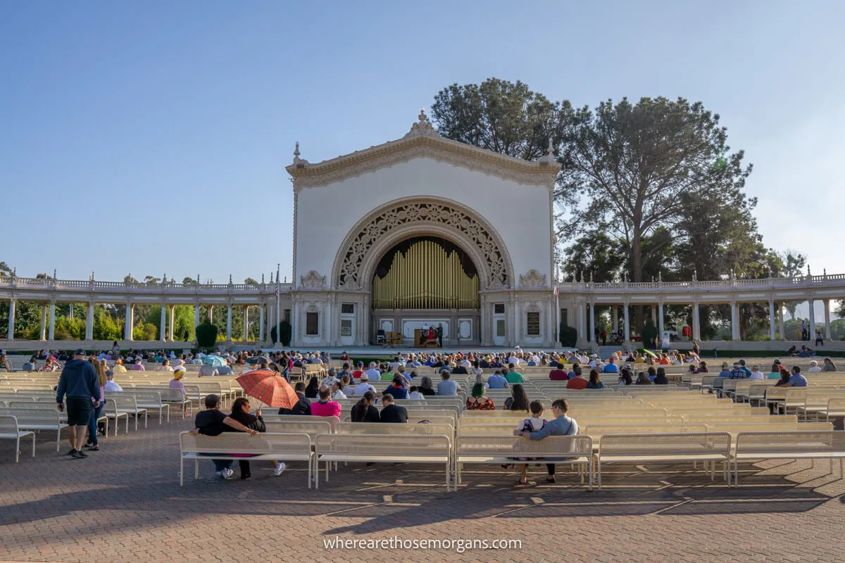 Amphitheater with people sat on long rows of benches leading to a large outdoor organ on a sunny day