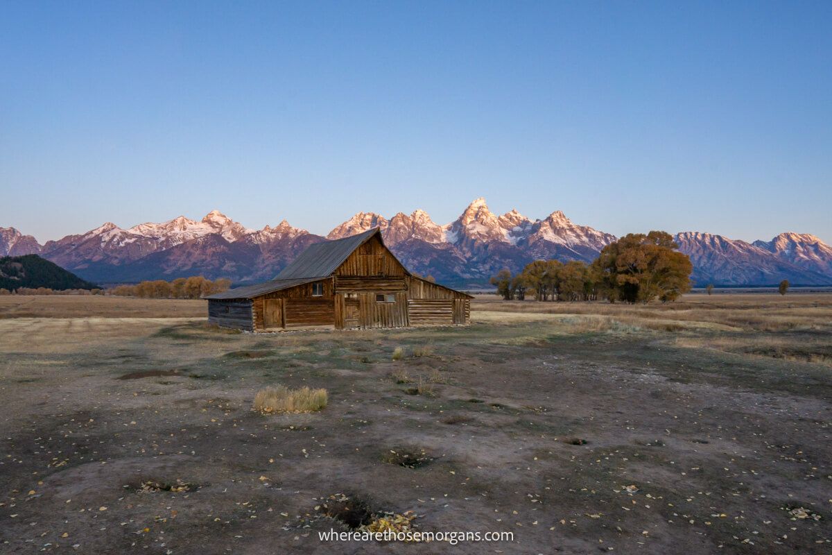 Vast flat landscape with grassland, a wooden building called T.A. Moulton Barn on Mormon Row and mountains in the background being lit up at sunrise