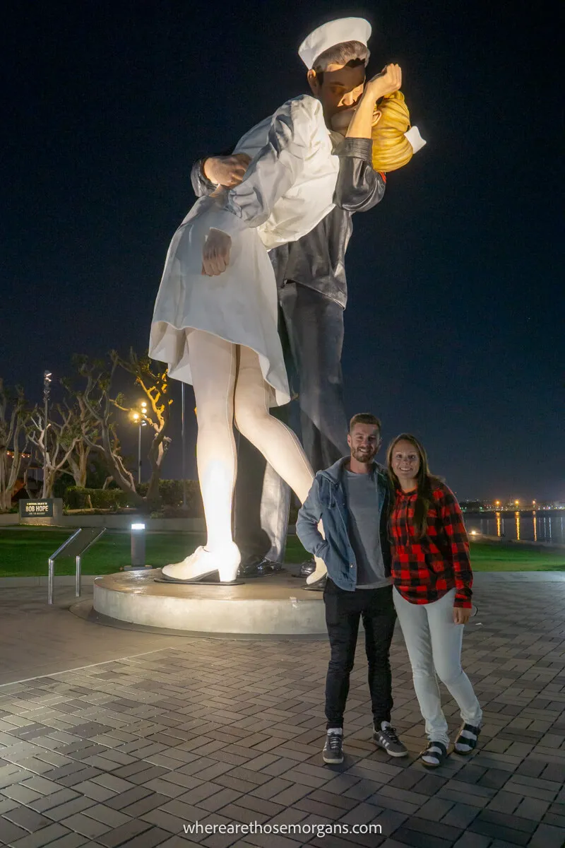 Couple standing together next to a large statue of a sailor kissing a local resident marking unconditional surrender in San Diego at night