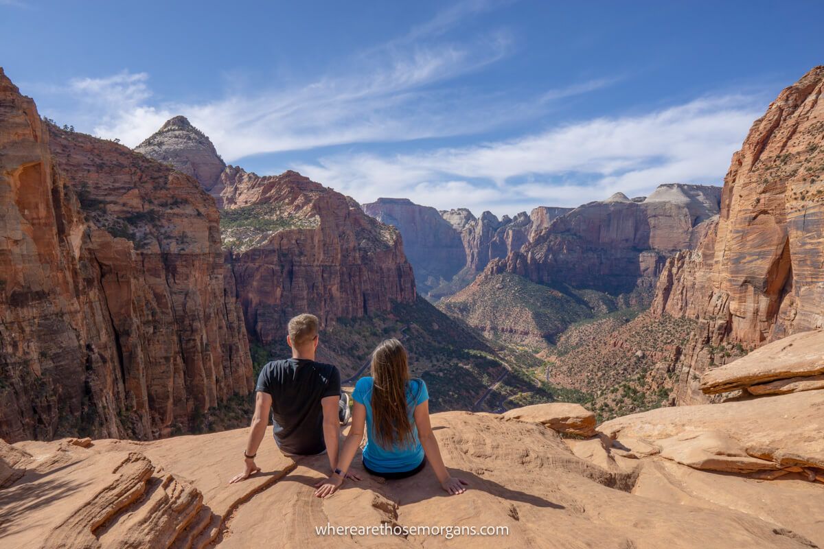 Two hikers sat together on a flat rocky surface looking out at a huge valley with tall cliff walls from Zion Canyon Overlook Trail in Utah on a sunny day